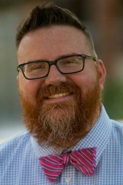 Portrait of Kevin Carmody, white male with short red hair and a full red beard. Kevin is wearing a blue checked shirt and a red/blue striped bowtie and is smiling.